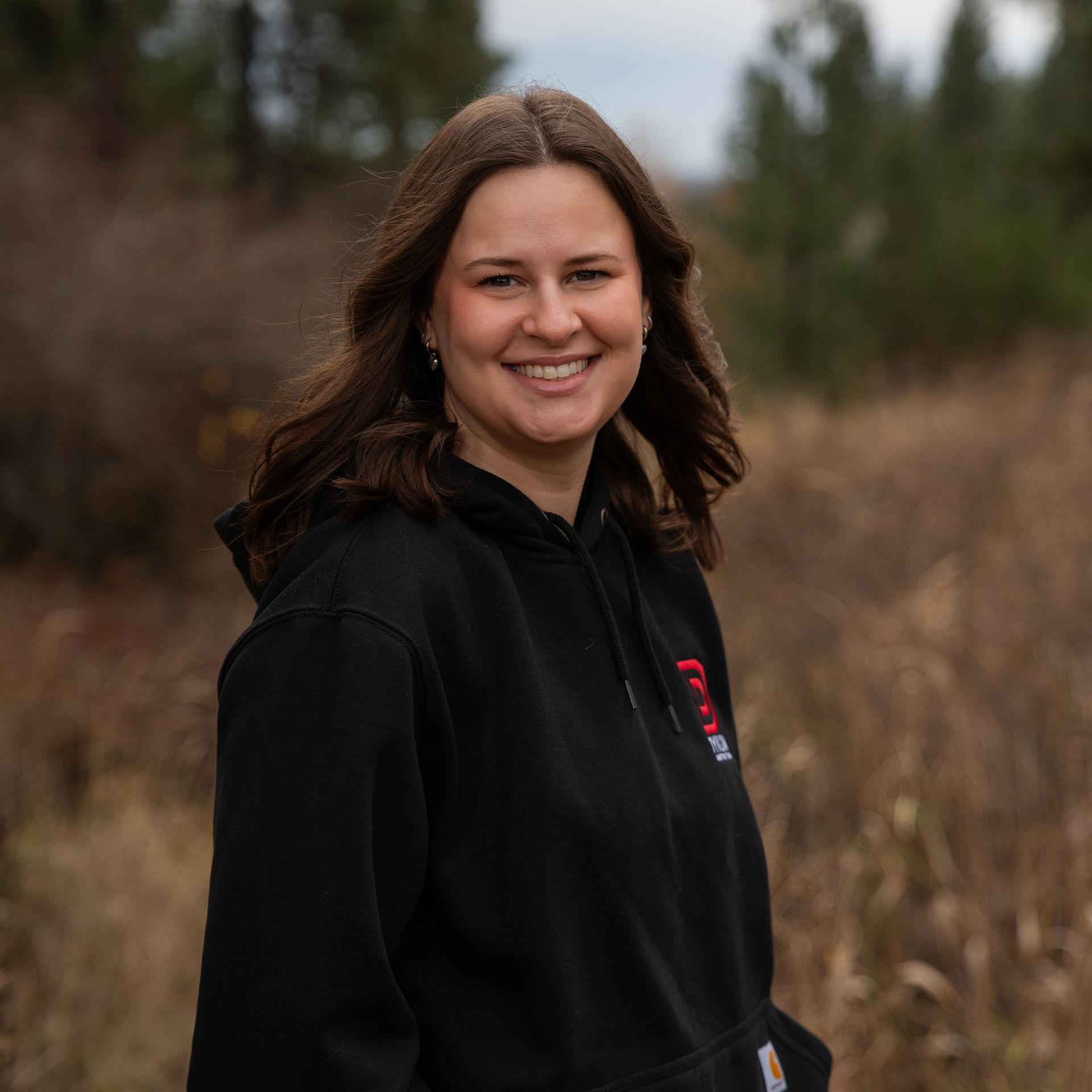 Smiling woman in a black hoodie standing outdoors in a grassy field with trees behind her