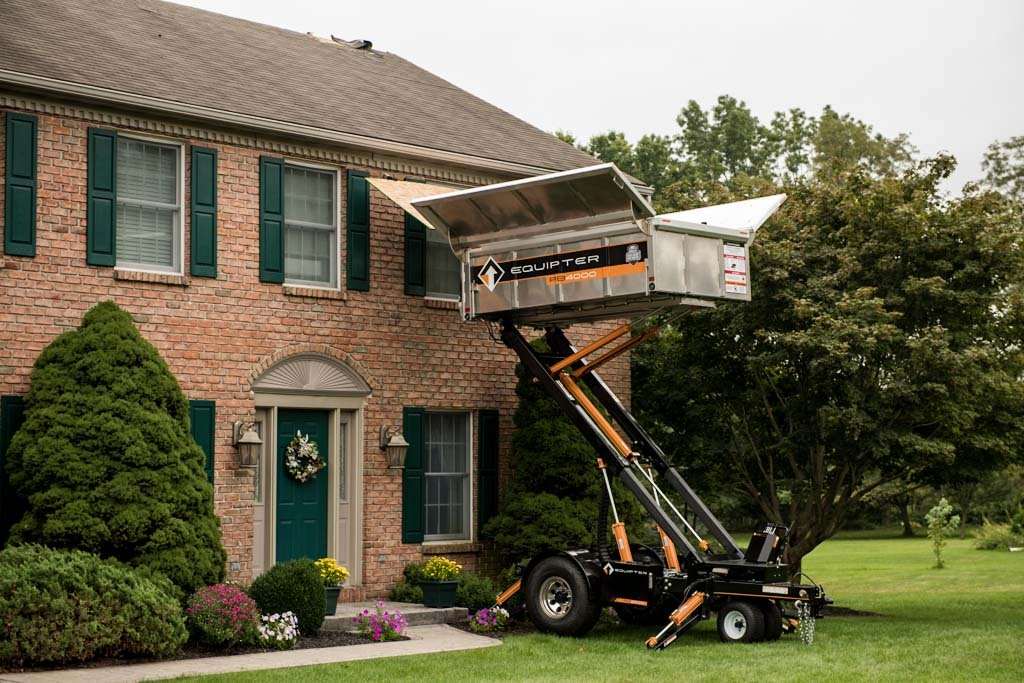 Worker in cherry picker trimming a second-story roofline on a brick house with green shutters.