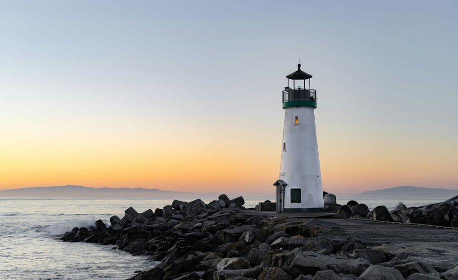 Lighthouse on a rocky breakwater at sunset with calm water and distant mountains.