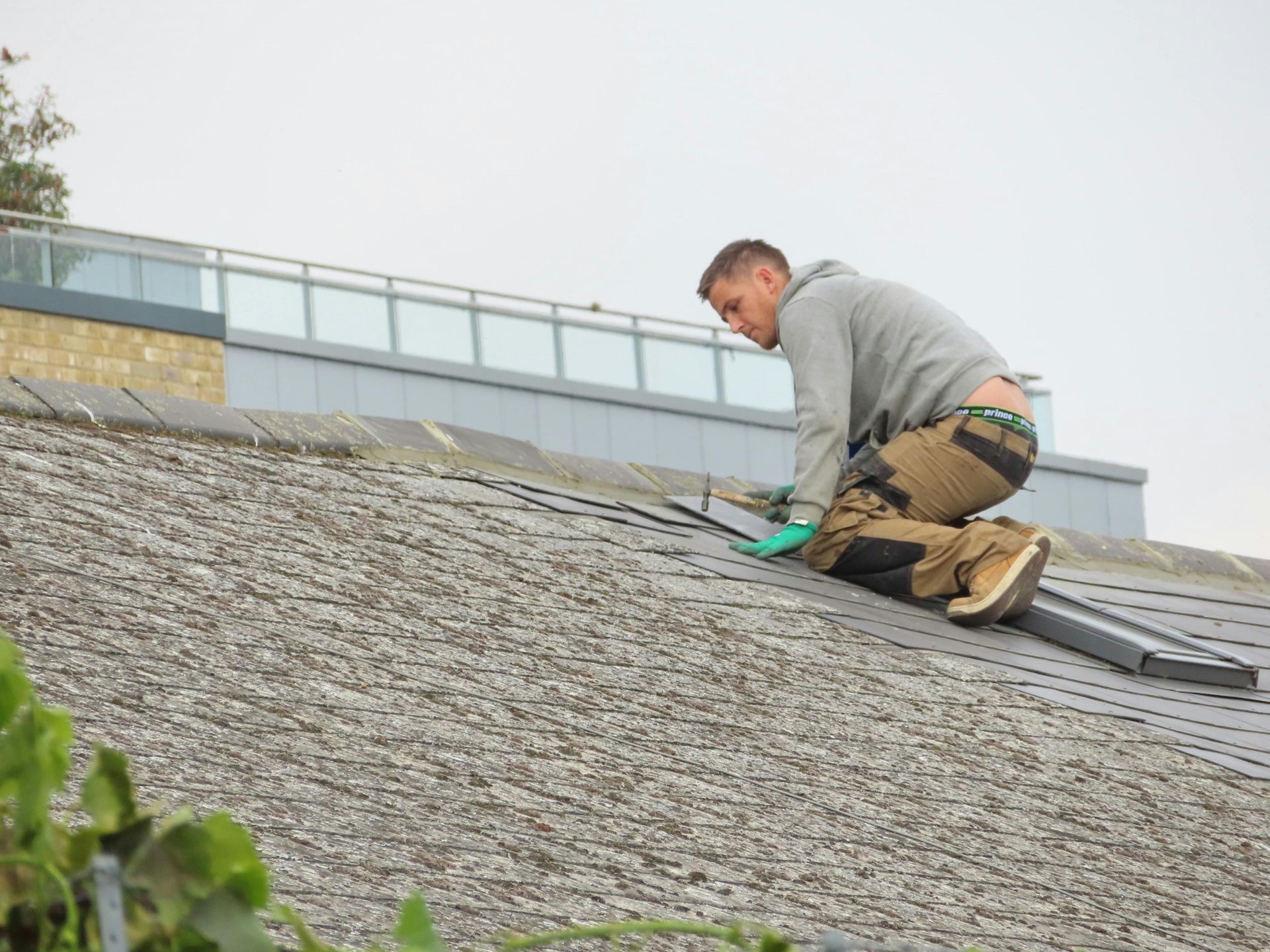 A person in work clothes kneels on a sloped shingled roof, installing long metal flashing.