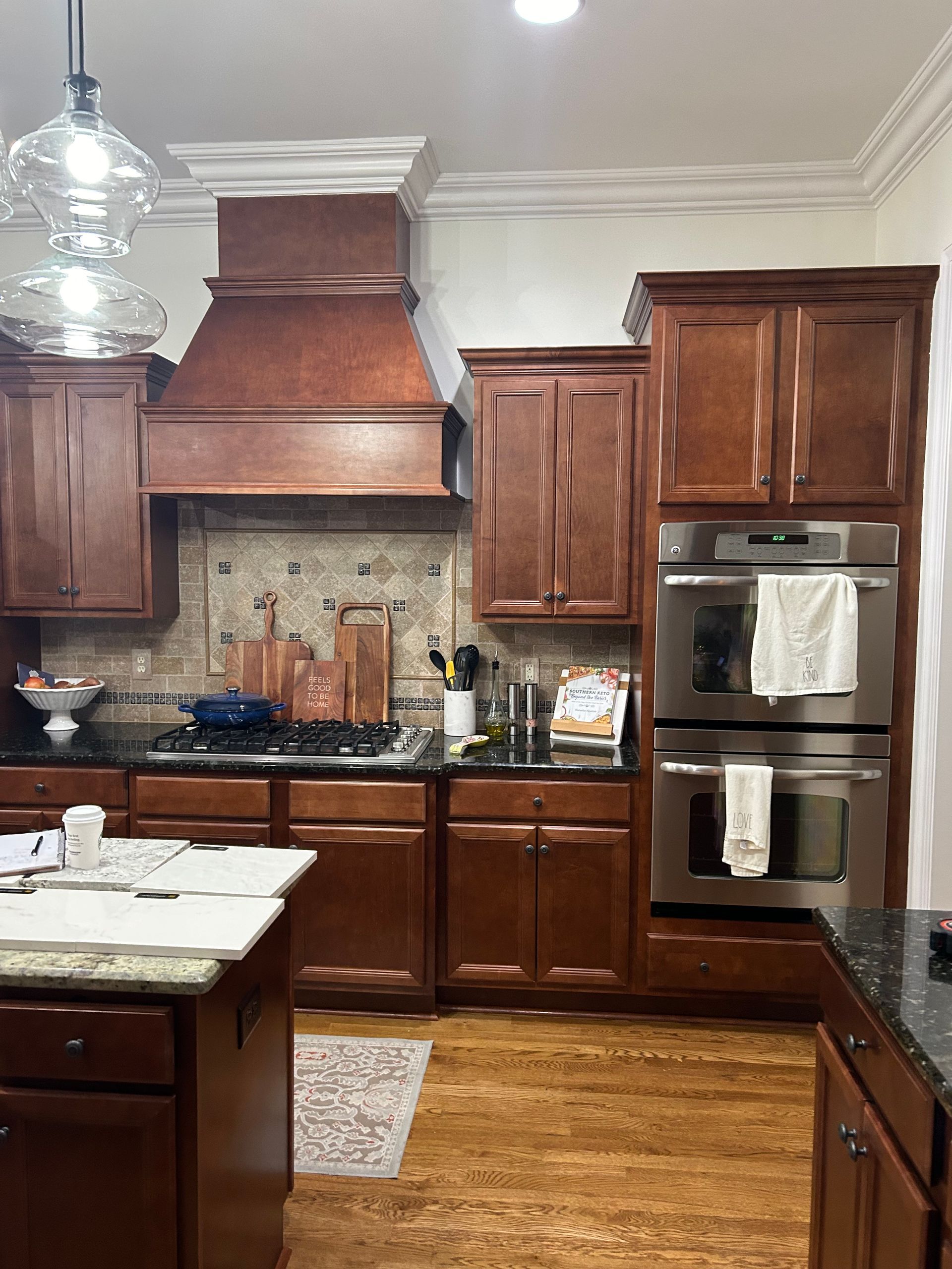 Small kitchen with white cabinets, island counter cluttered with items, and open doorway to a living room.