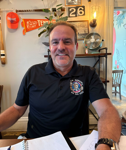 Smiling man in a black polo shirt seated at a table in a cozy restaurant or bar setting.