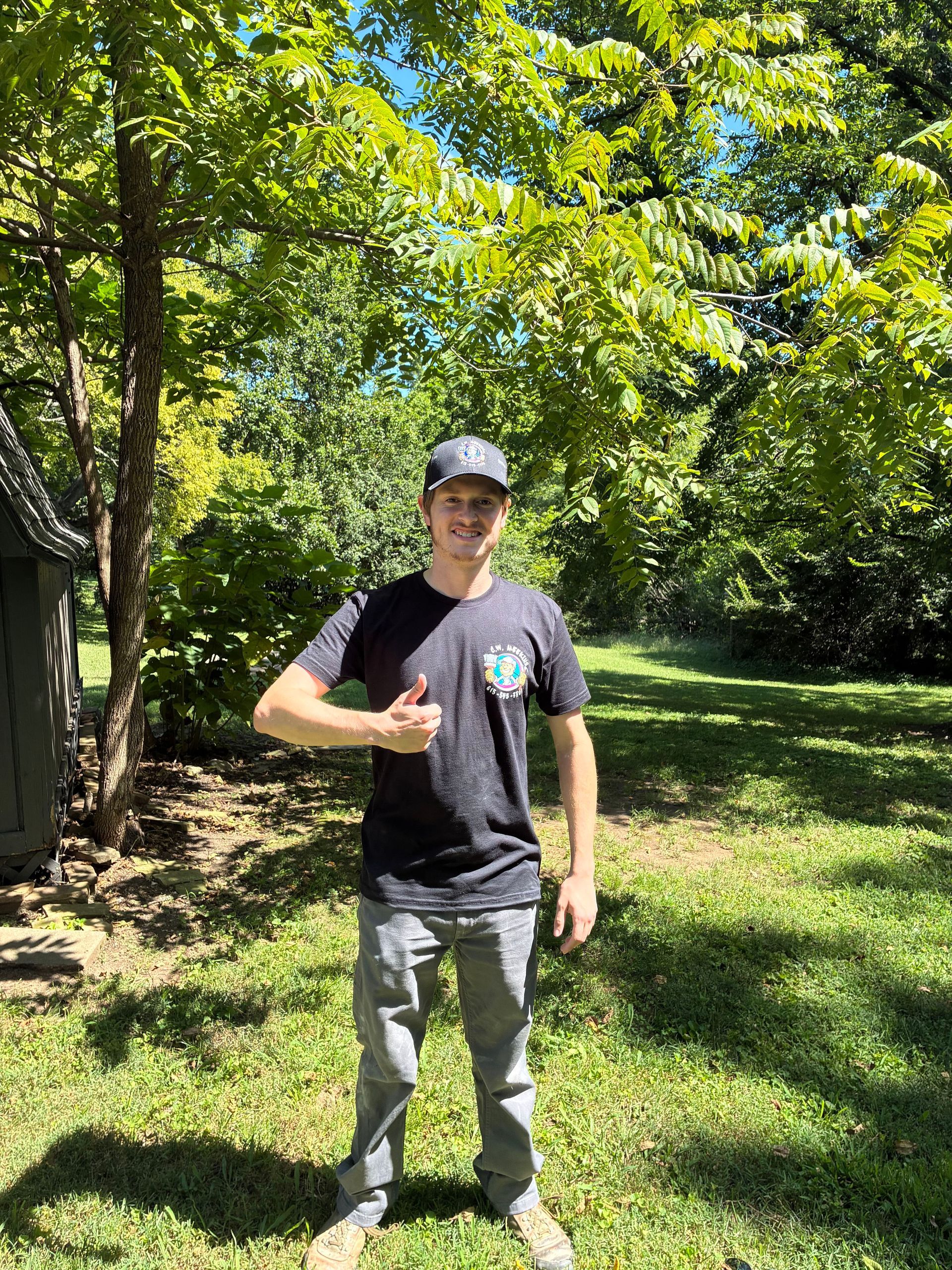 Person standing on a grassy lawn giving a thumbs-up in a sunny, tree-filled yard