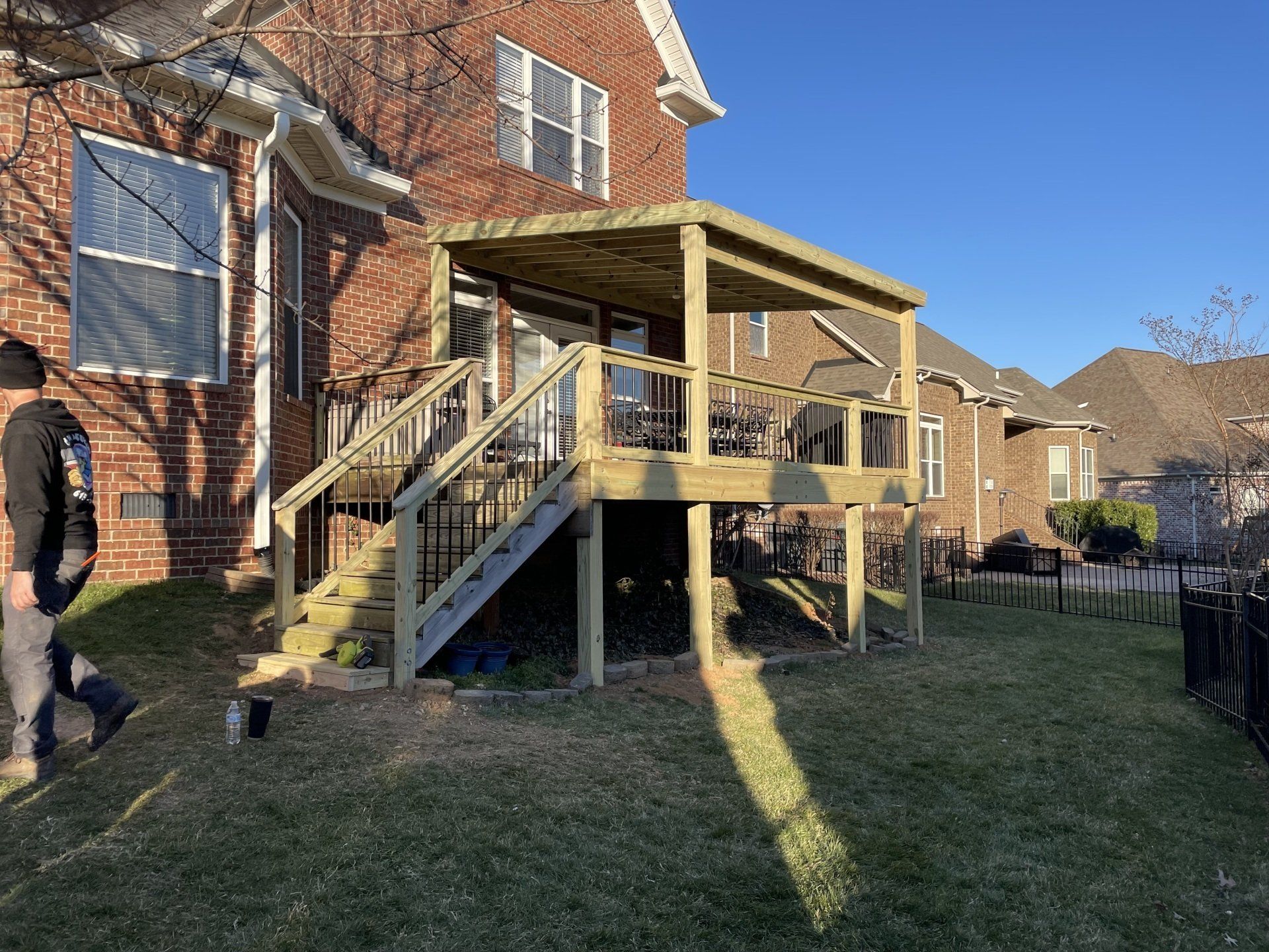 Wooden ramp and deck attached to brick house in a sunny backyard; person walking at left.
