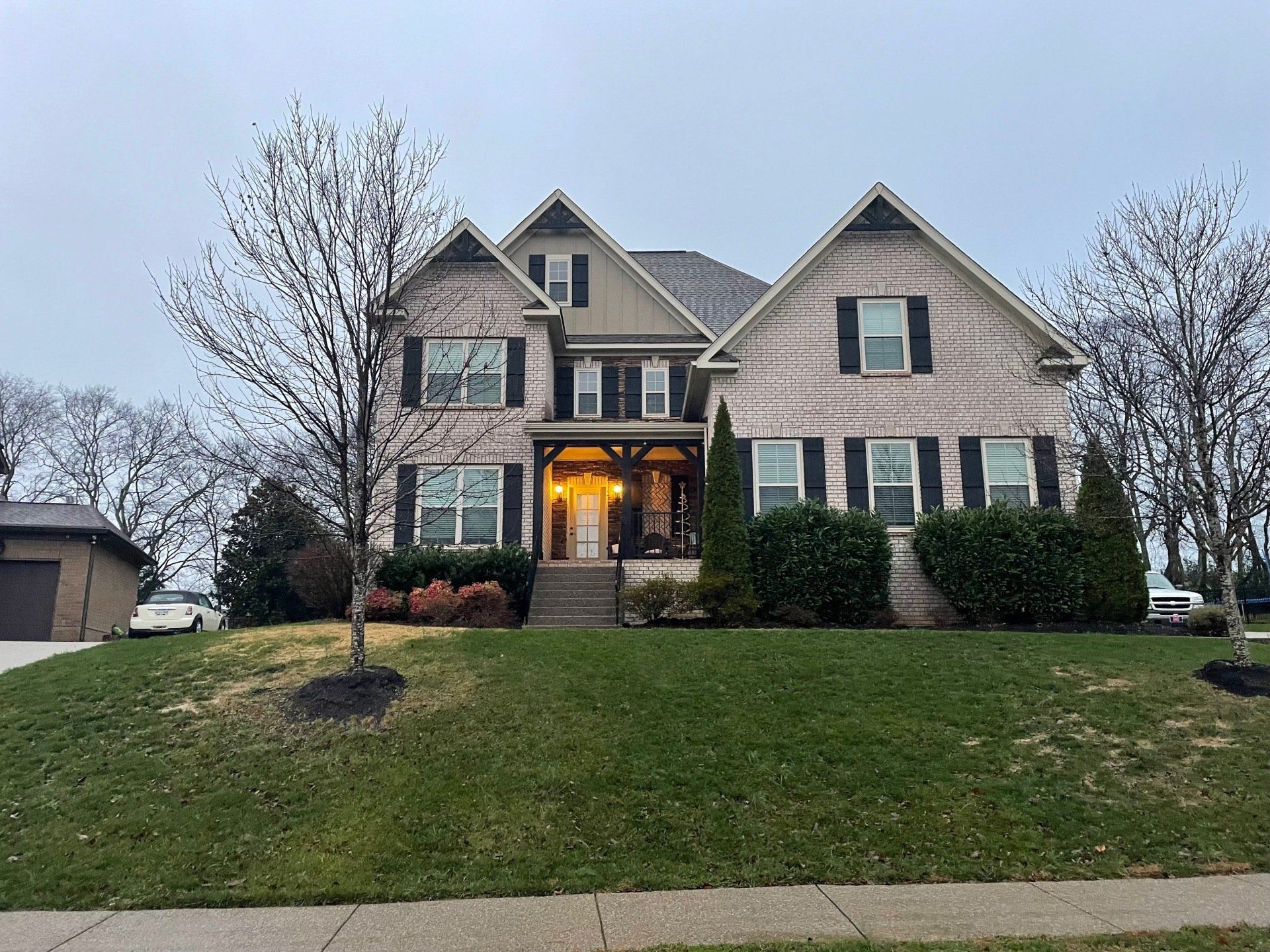 Suburban two-story house with lit porch, gray siding, dark shutters, and a green front lawn