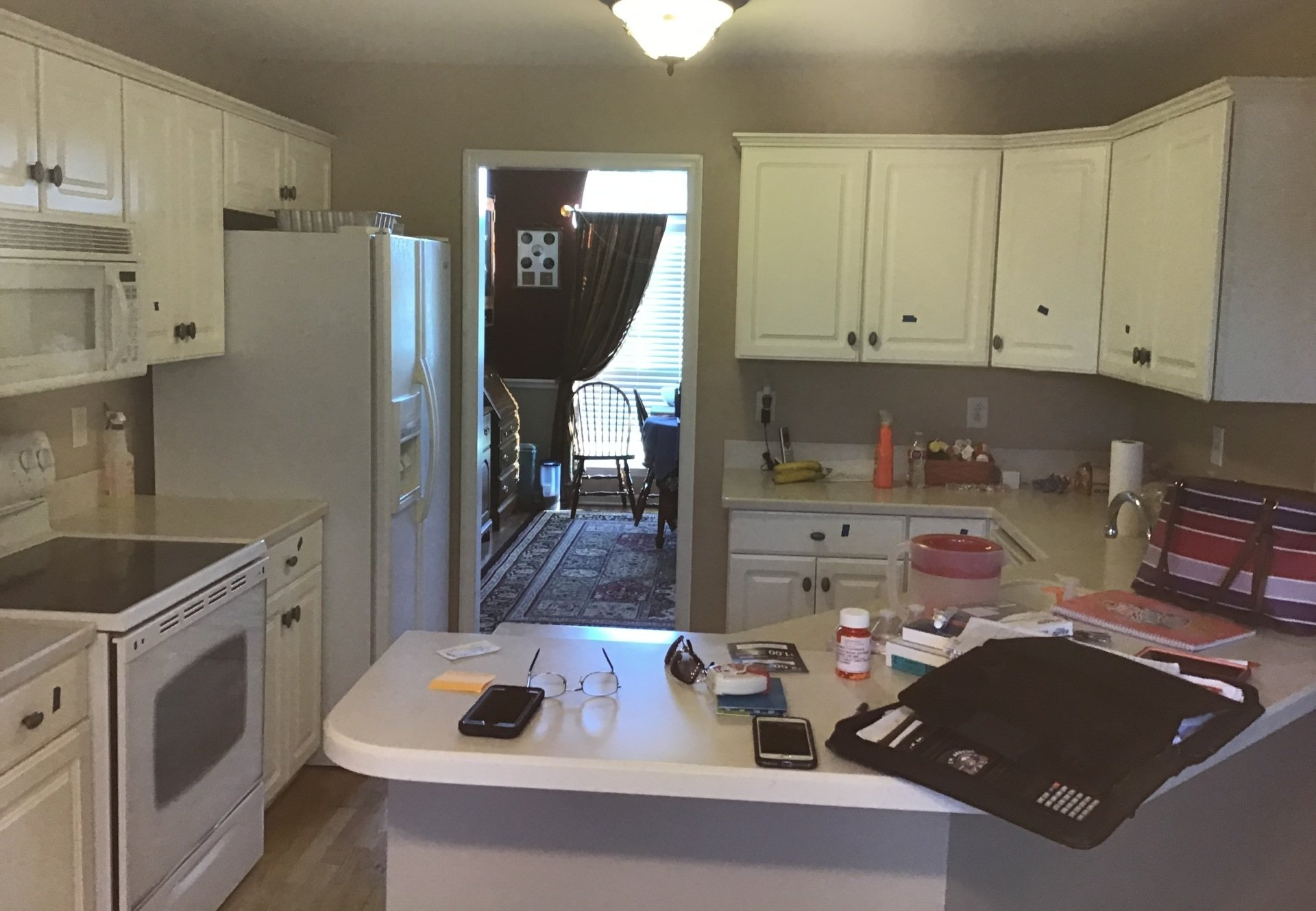 Cluttered white kitchen with cabinets, stove, refrigerator, and a central island holding scattered items.