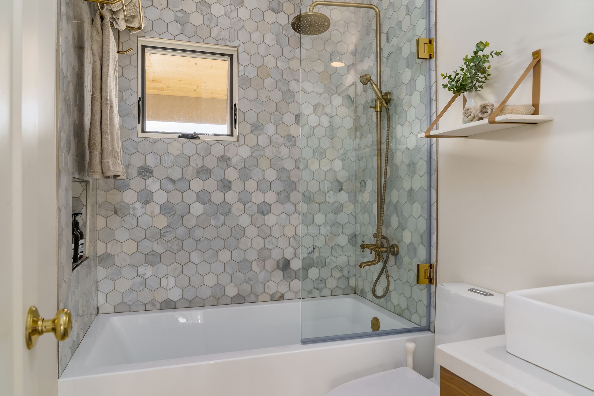Bright bathroom with marble-tile shower, glass door, tub, and white vanity with a plant on shelf.