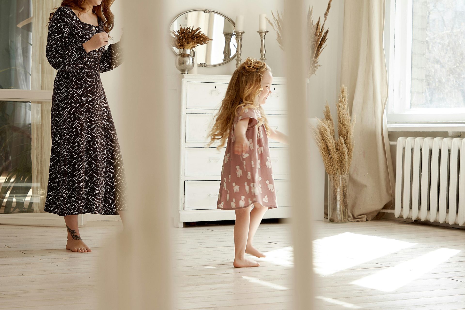 Woman in dark dress stands near a barefoot child in a pink dress dancing in a sunlit room.