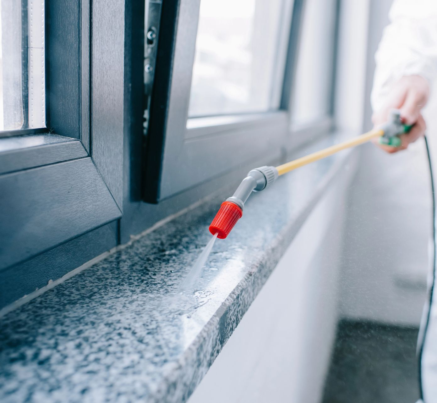 Person cleaning a windowsill with a spray nozzle, misting water along the gray ledge indoors