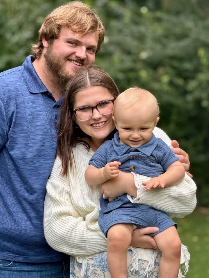 Smiling family of three outdoors, with parents holding a baby in blue overalls.