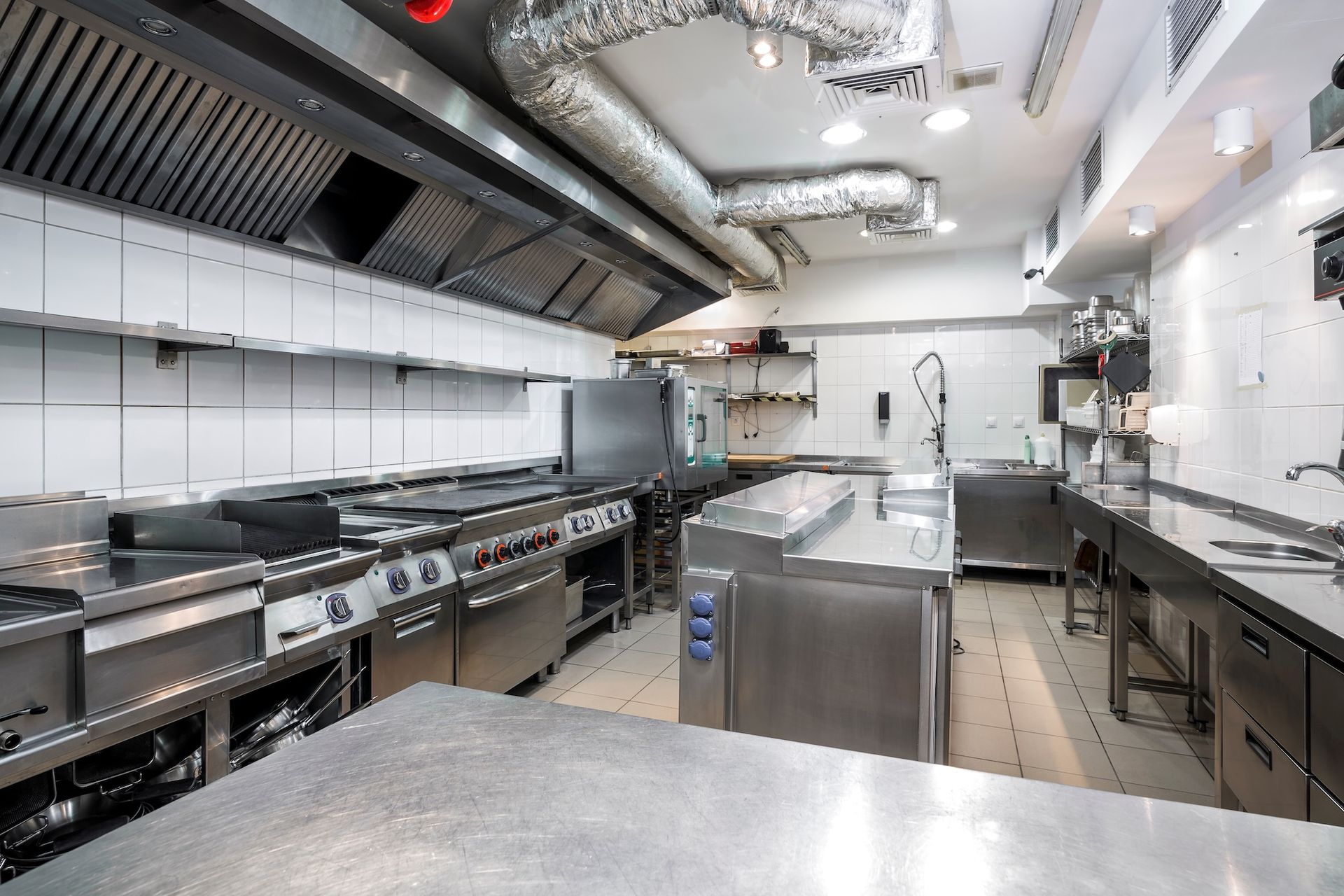 Commercial stainless steel restaurant kitchen with stoves, prep tables, and overhead ventilation