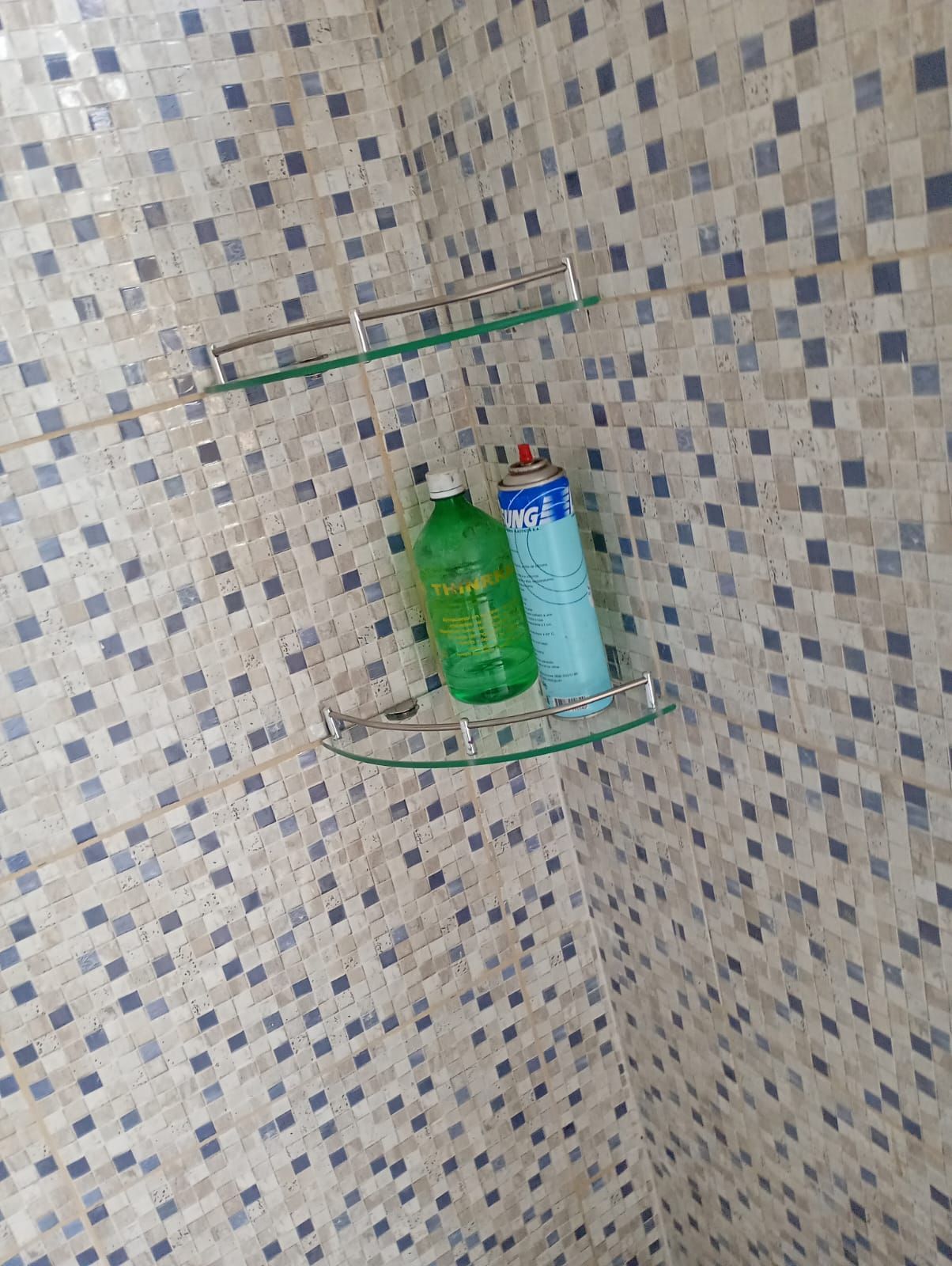 Rincón de ducha con pared de azulejos de mosaico, estante de vidrio y dos botellas de jabón o champú.