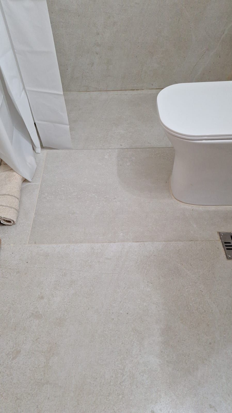 Suelo del baño con azulejos beige, borde de la cortina de la ducha y parte de un inodoro blanco.