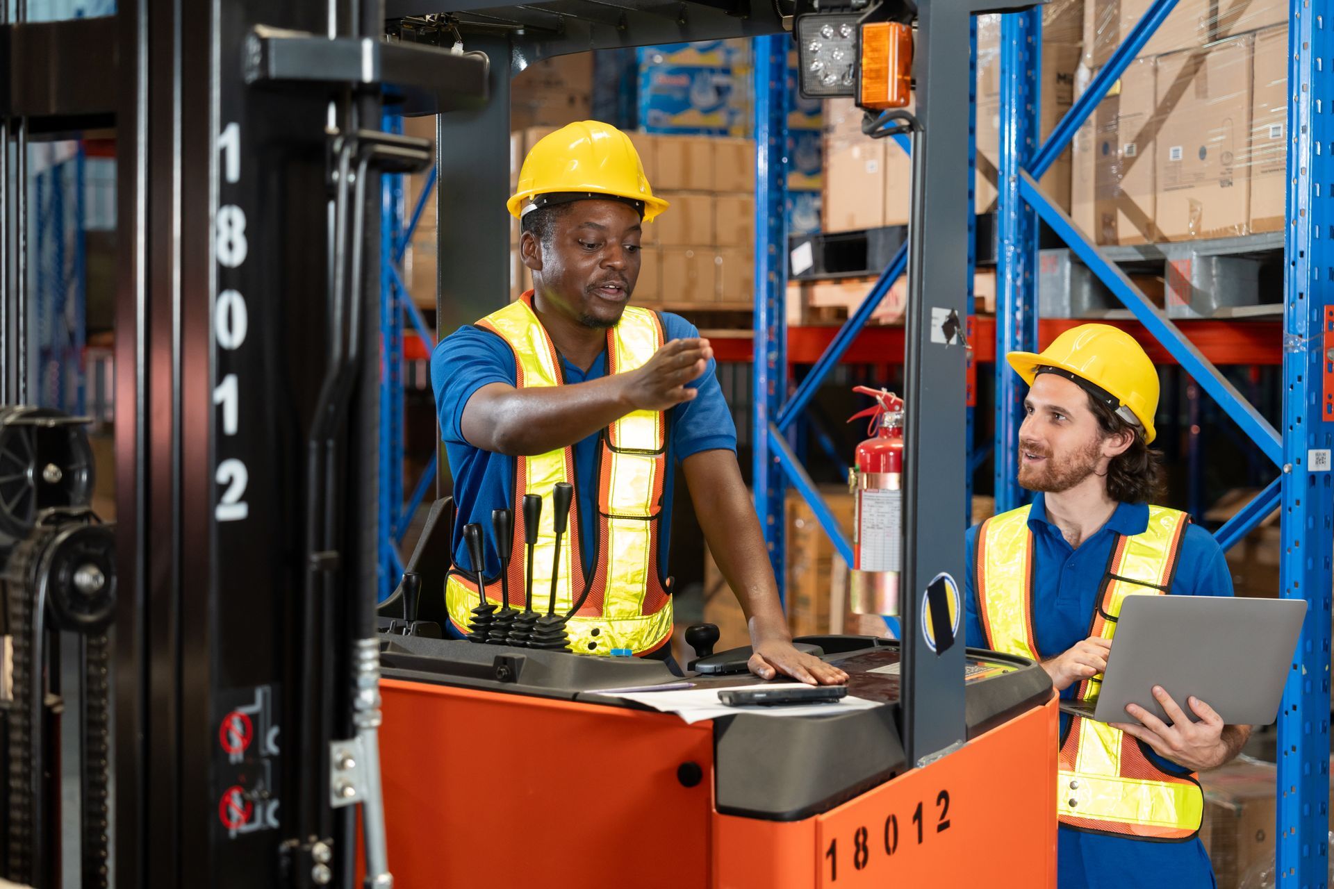 Two warehouse workers in hard hats inspecting boxes on a forklift in a storage aisle