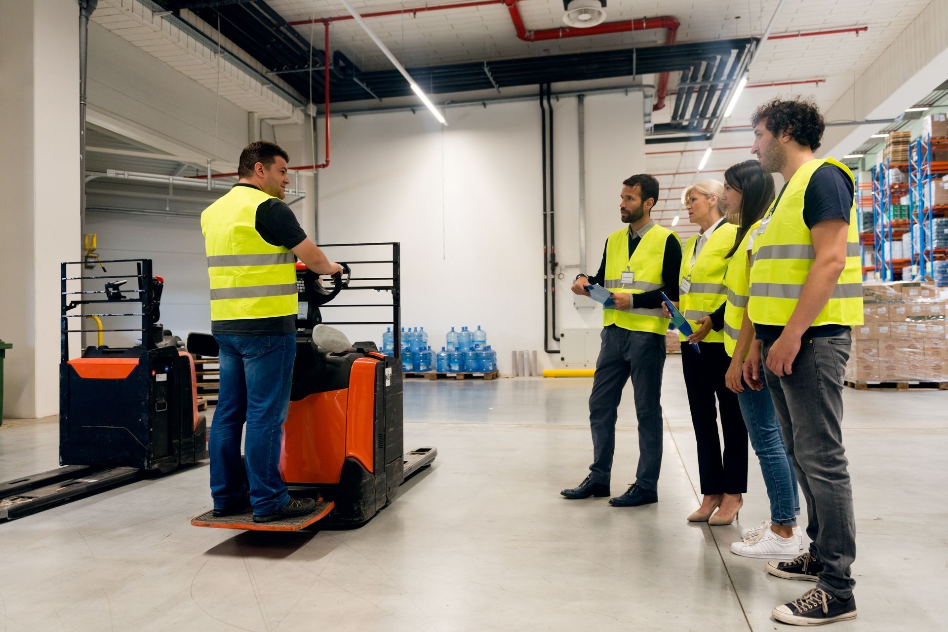 Workers in safety vests listening to a forklift operator in a warehouse.