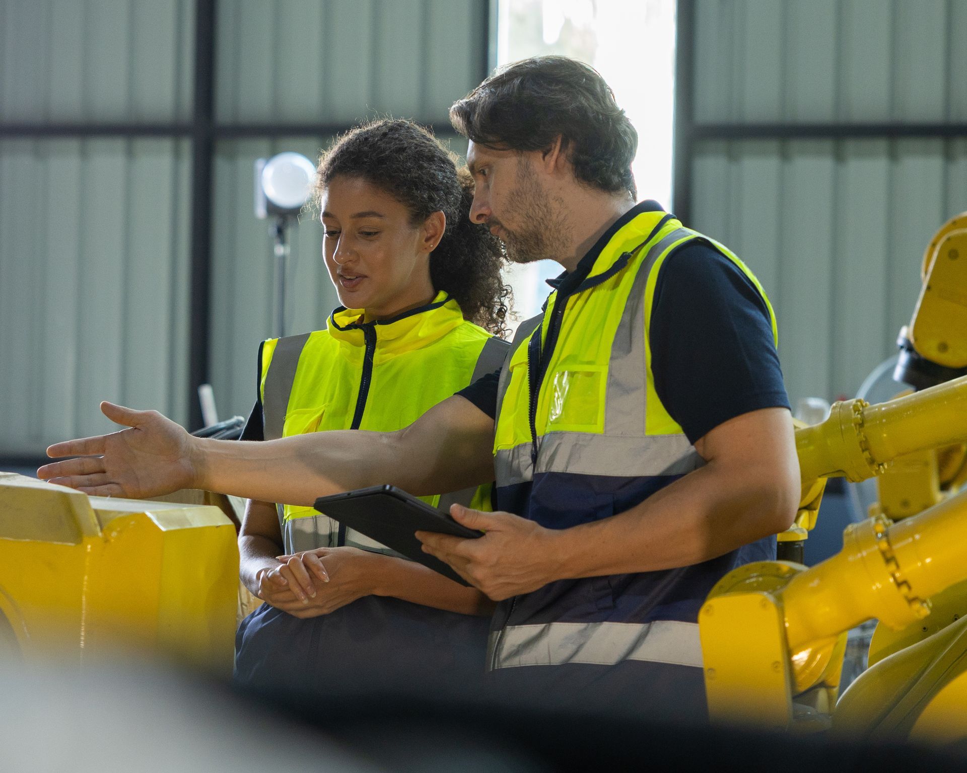 Two workers in yellow safety vests review a tablet beside industrial machinery.