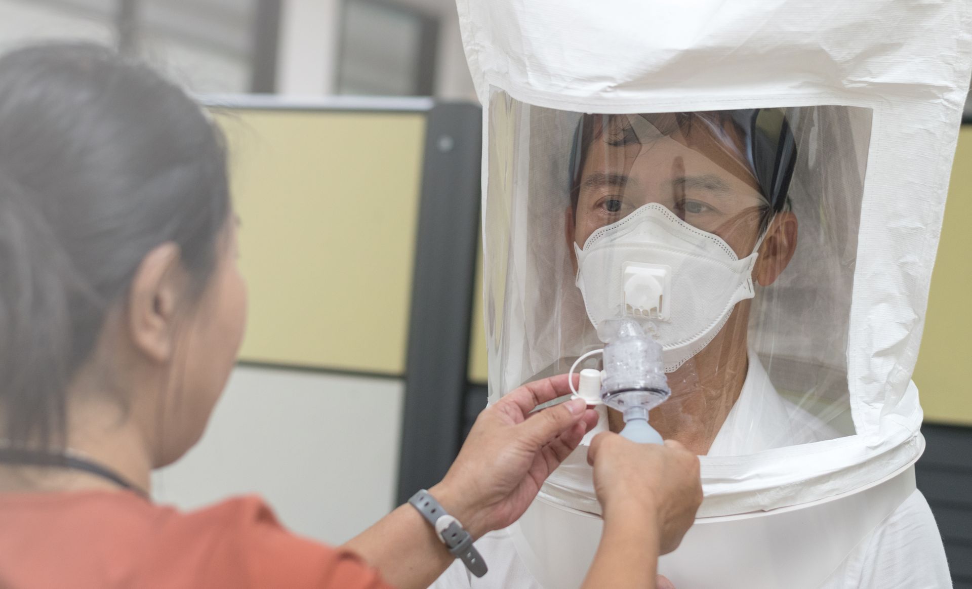 Person adjusting a face mask inside a white isolation hood with a clear front panel