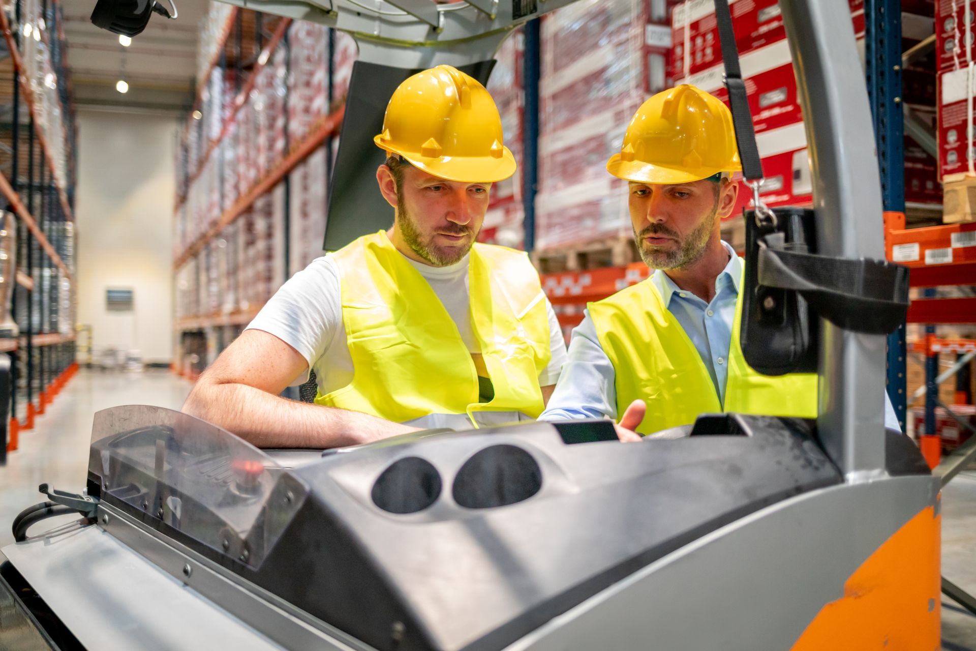 Two warehouse workers in yellow hard hats and vests review a forklift control panel in an aisle of shelves.