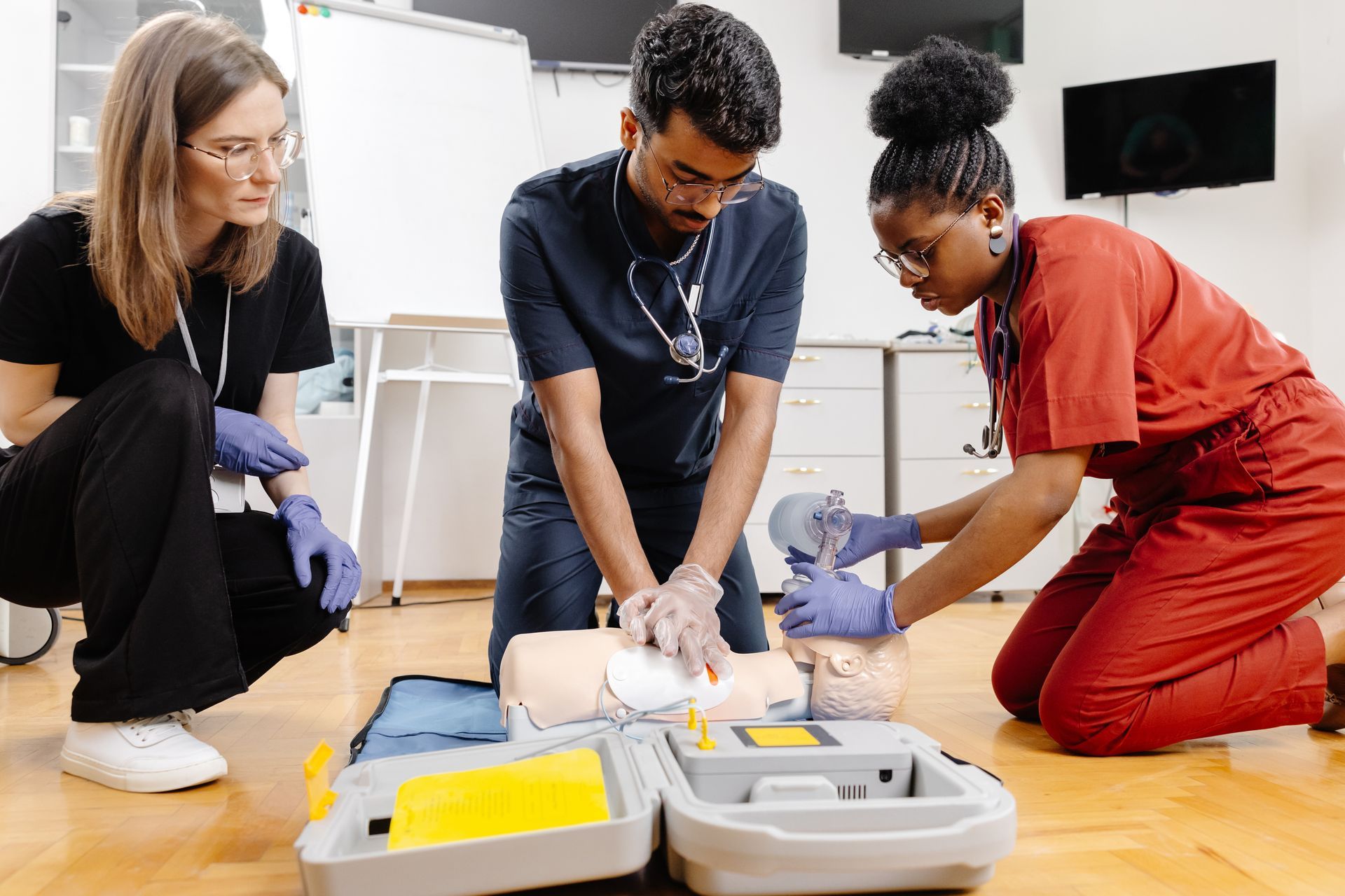 Three people kneel on a gym floor, practicing CPR on a training manikin with first-aid kits nearby.