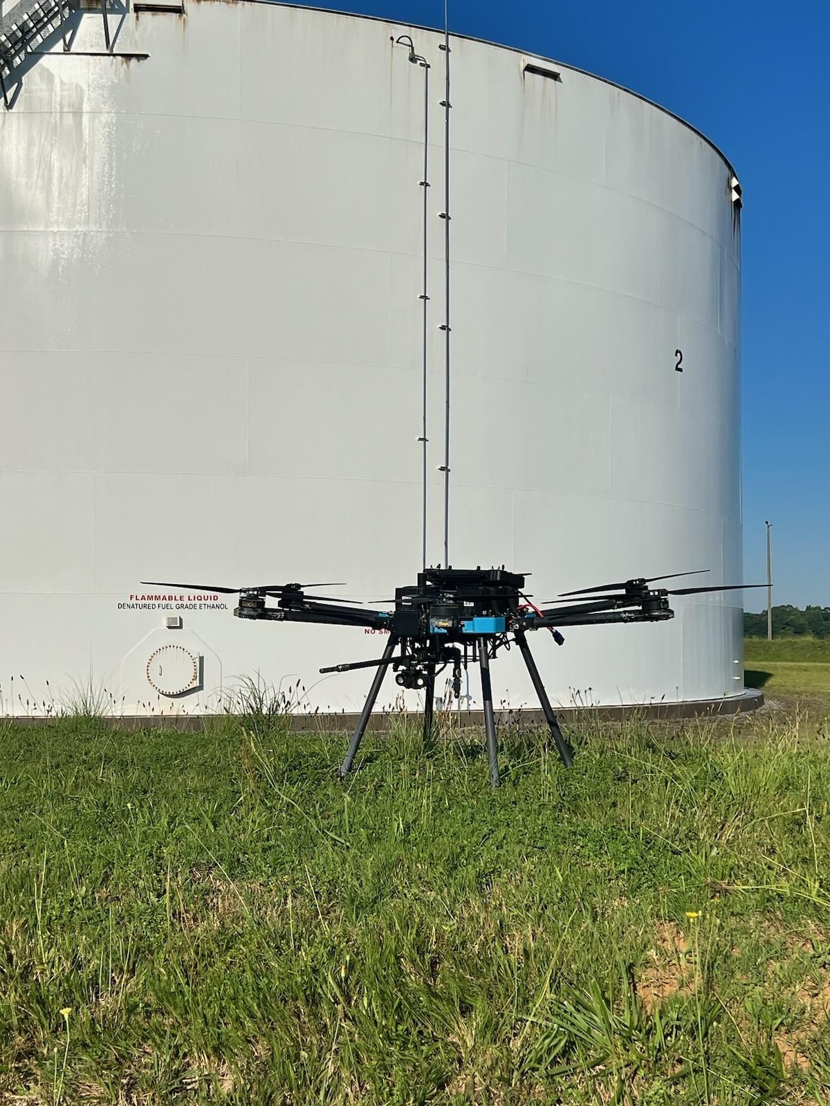 Black drone on grass in front of a large white storage tank under a blue sky