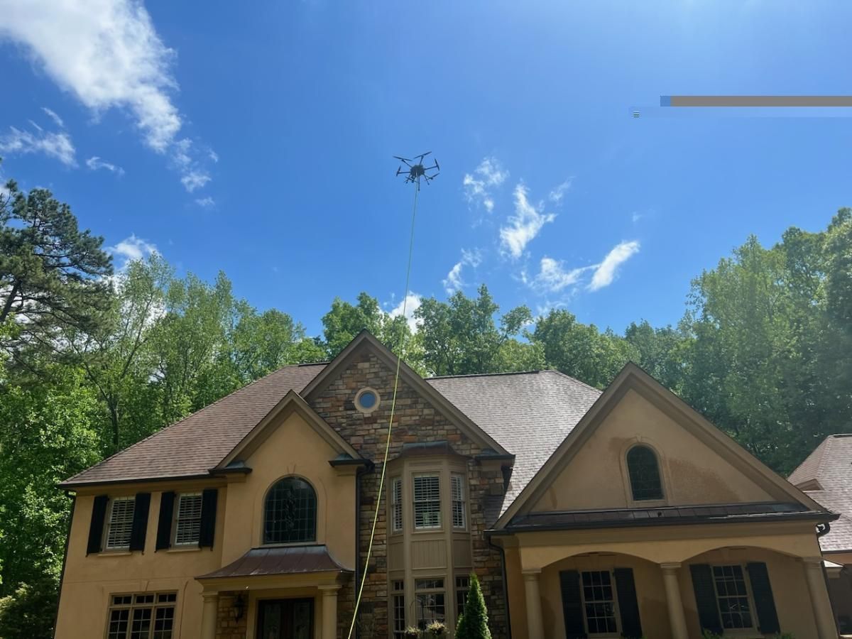 Large tan house with a drone flying overhead under a blue sky, surrounded by trees