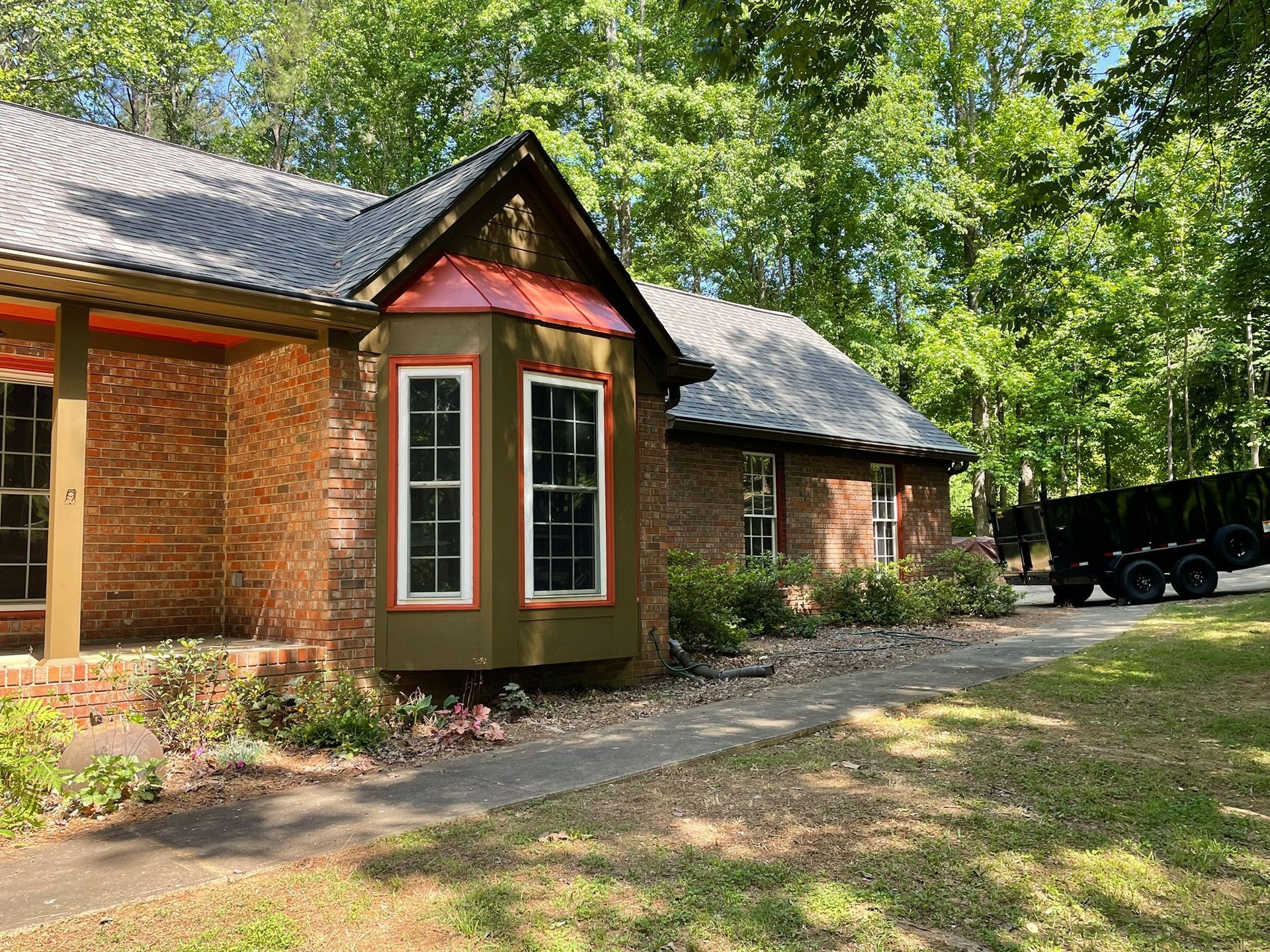 Brick house with a bay window and black carport, surrounded by trees and a dirt driveway