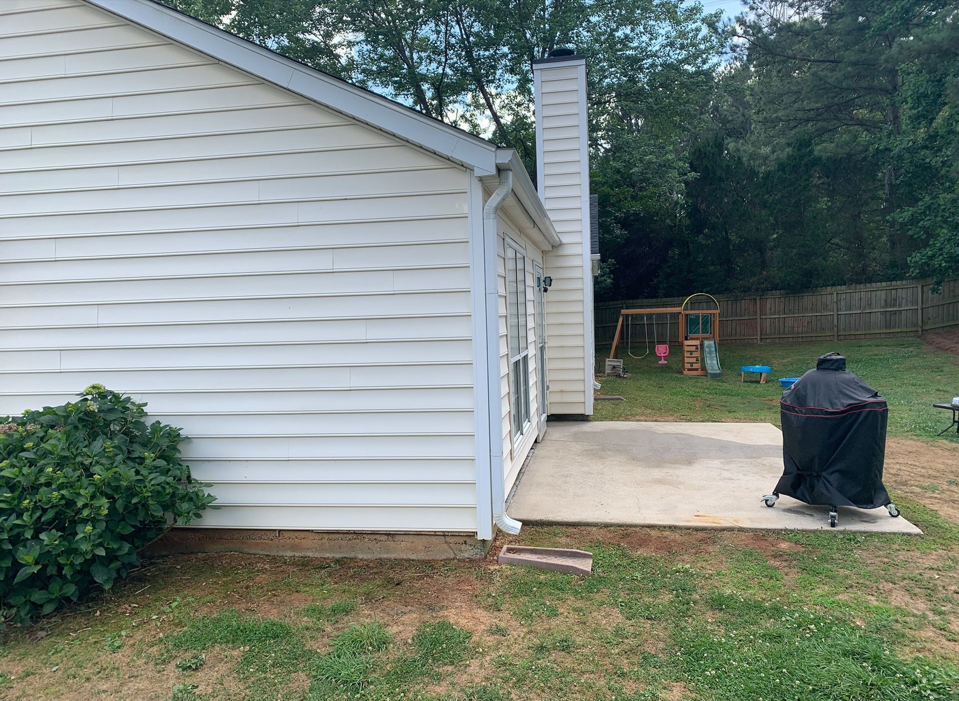 Side view of a white house with a concrete patio, grill, and backyard playset under trees.