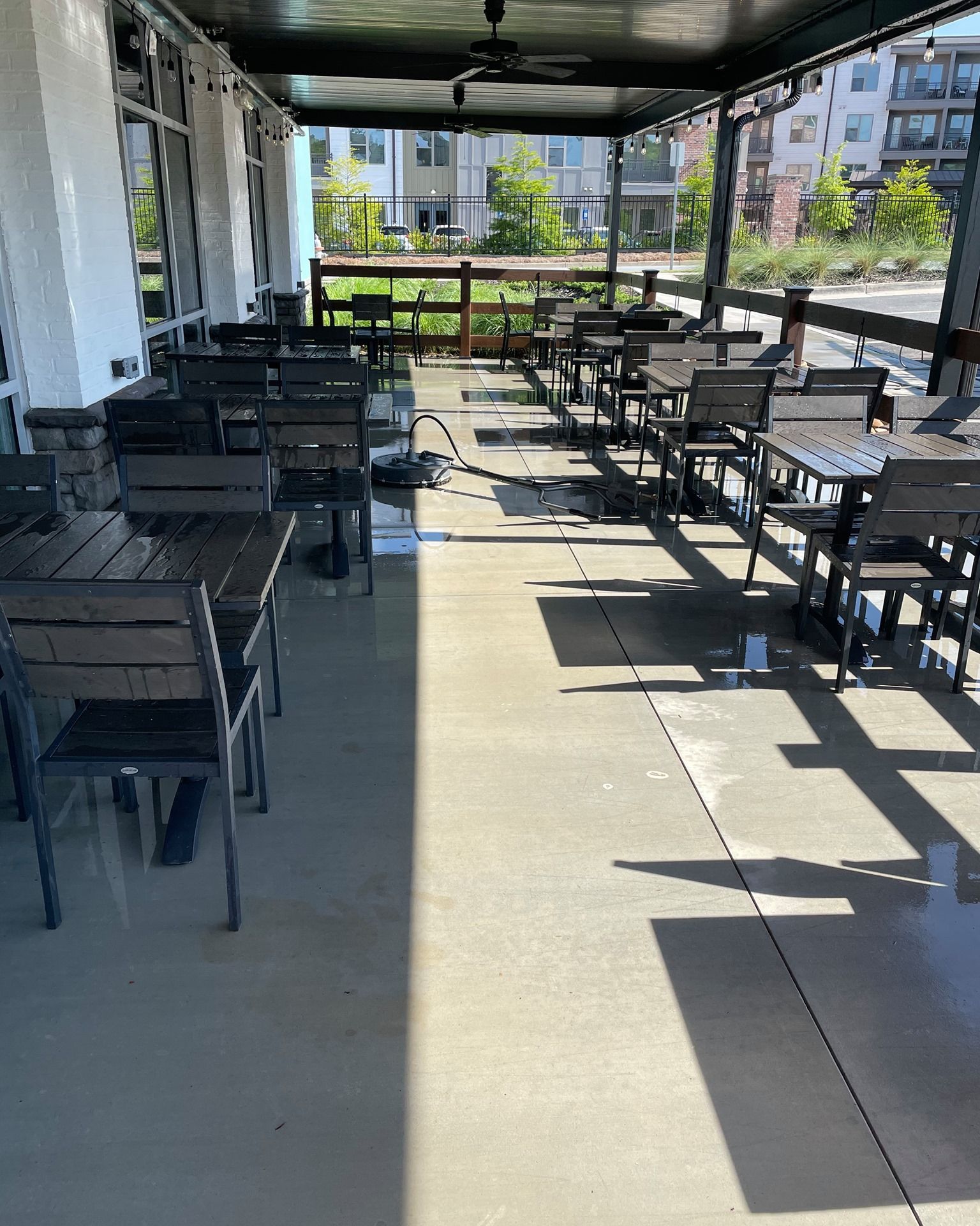 Covered outdoor patio with black tables and chairs, sunlight casting shadows, overlooking a courtyard.