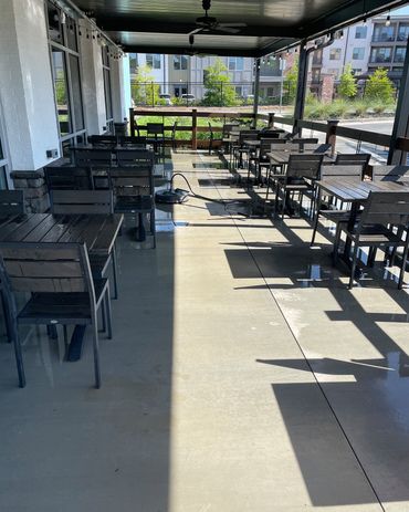 Empty outdoor patio with black tables and chairs under a covered awning, in bright sunlight.