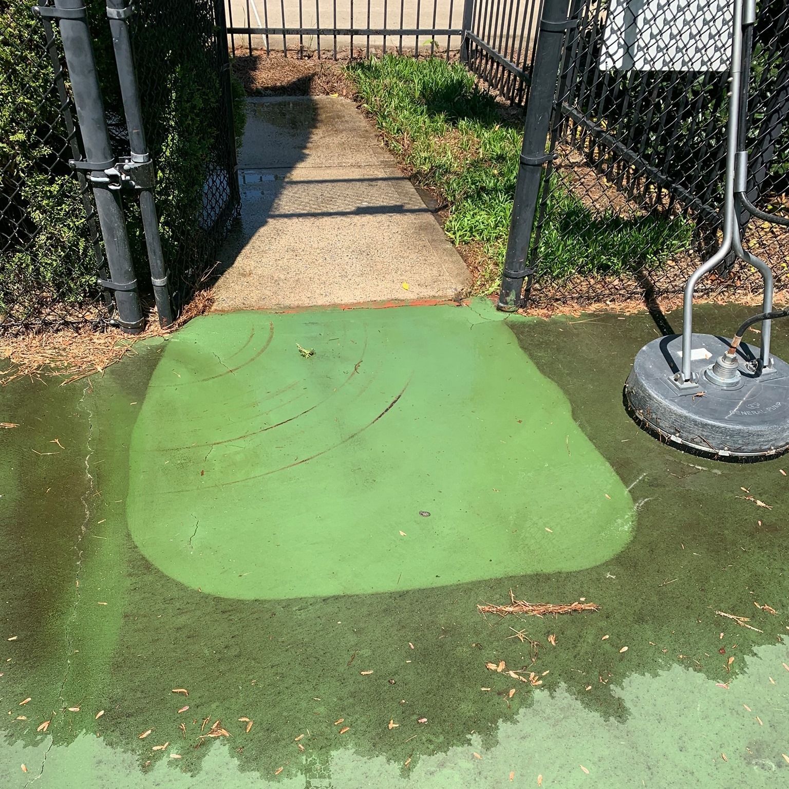 Green painted patch on a sidewalk entrance beside a chain-link fence and metal post.