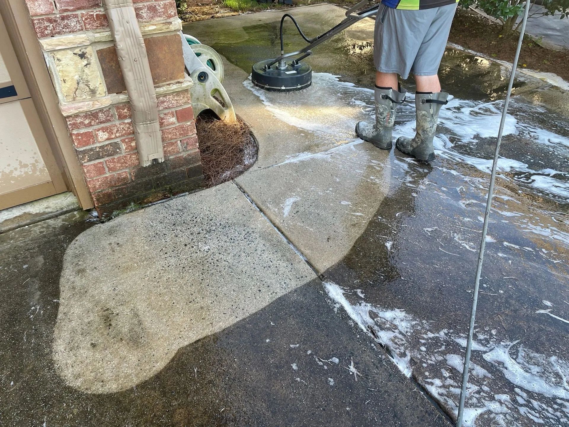 Person power-washing a concrete patio beside a brick house, with soapy water spraying across the ground