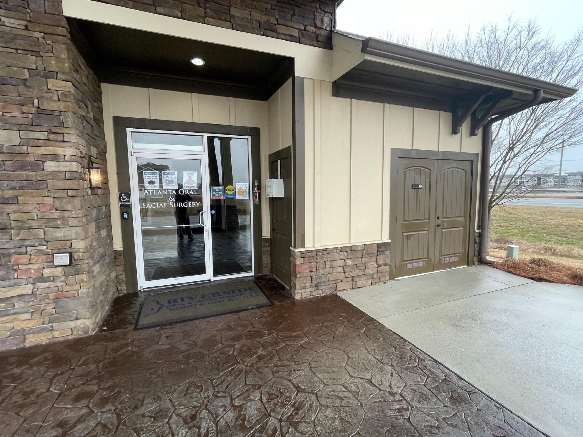 Building entrance with glass doors, stone facade, and wet pavement beside a concrete walkway