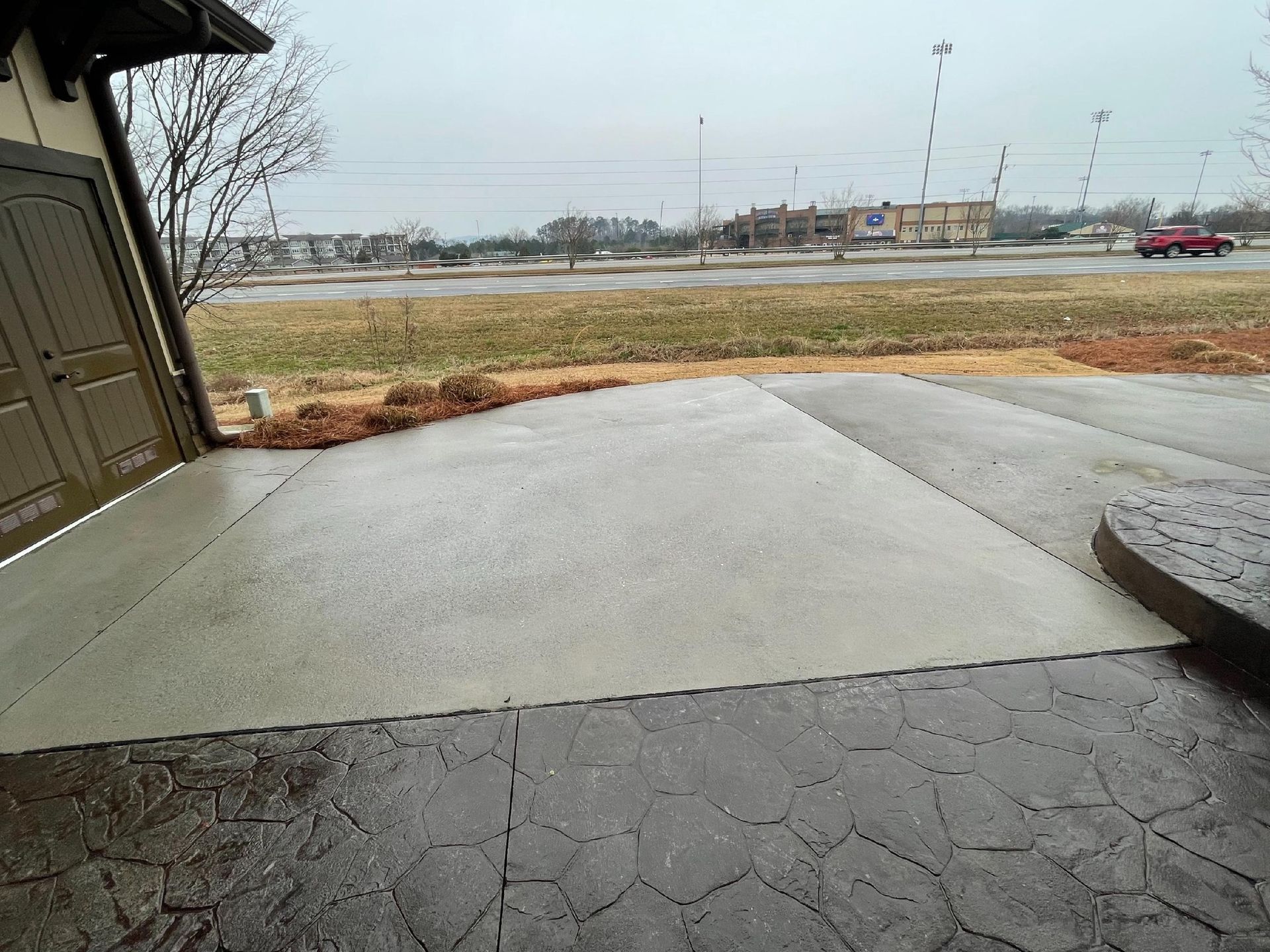 Concrete driveway leading to a road, with grass, overcast sky, and a parked red car in the distance