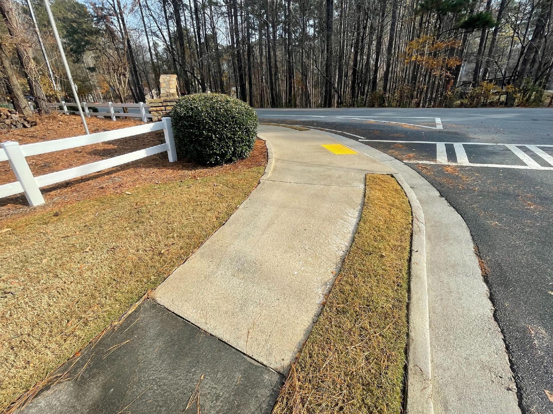 Curved concrete sidewalk ramps to a road crossing beside a white fence and autumn trees.