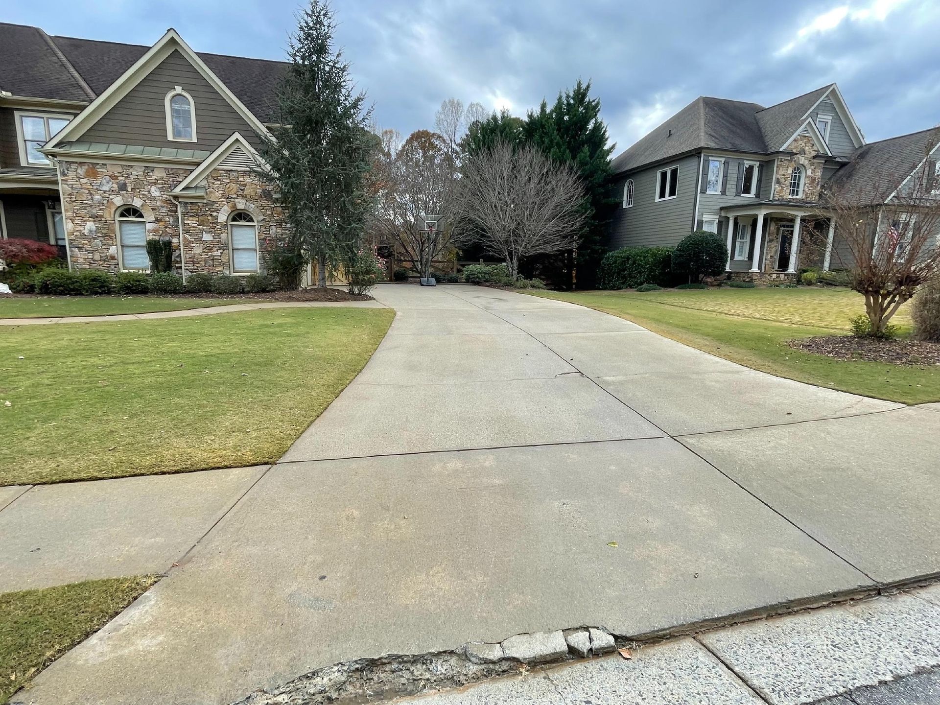 Stone suburban homes with a long concrete driveway and green lawns in front of wooded trees
