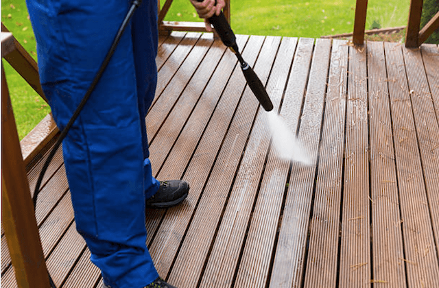Person pressure-washing a wooden deck with a black wand, wearing blue pants and black shoes.