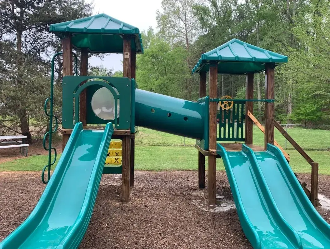 Children’s playground with two teal slides, connecting tunnel, and wood-chip ground under trees