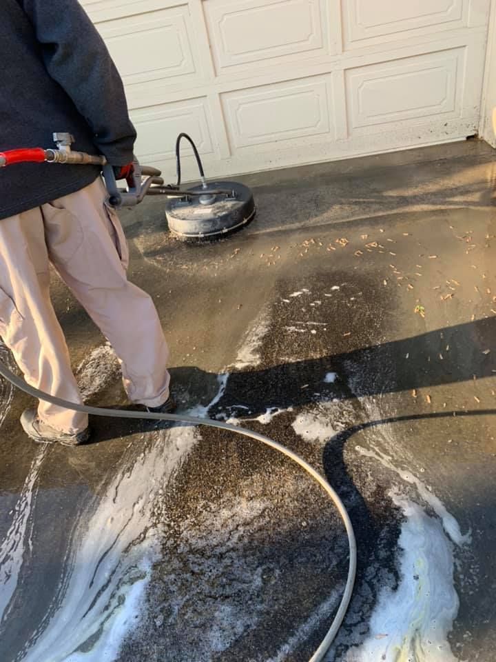 Person power-washing a driveway with soap and hose near a garage door.