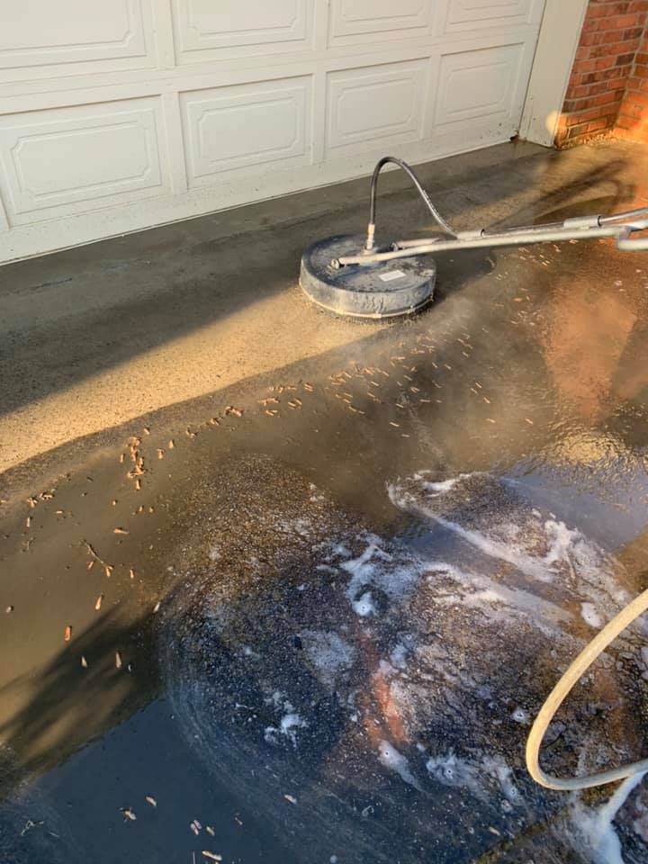 Person pressure-washing a driveway beside a garage, with white foam and wet pavement visible