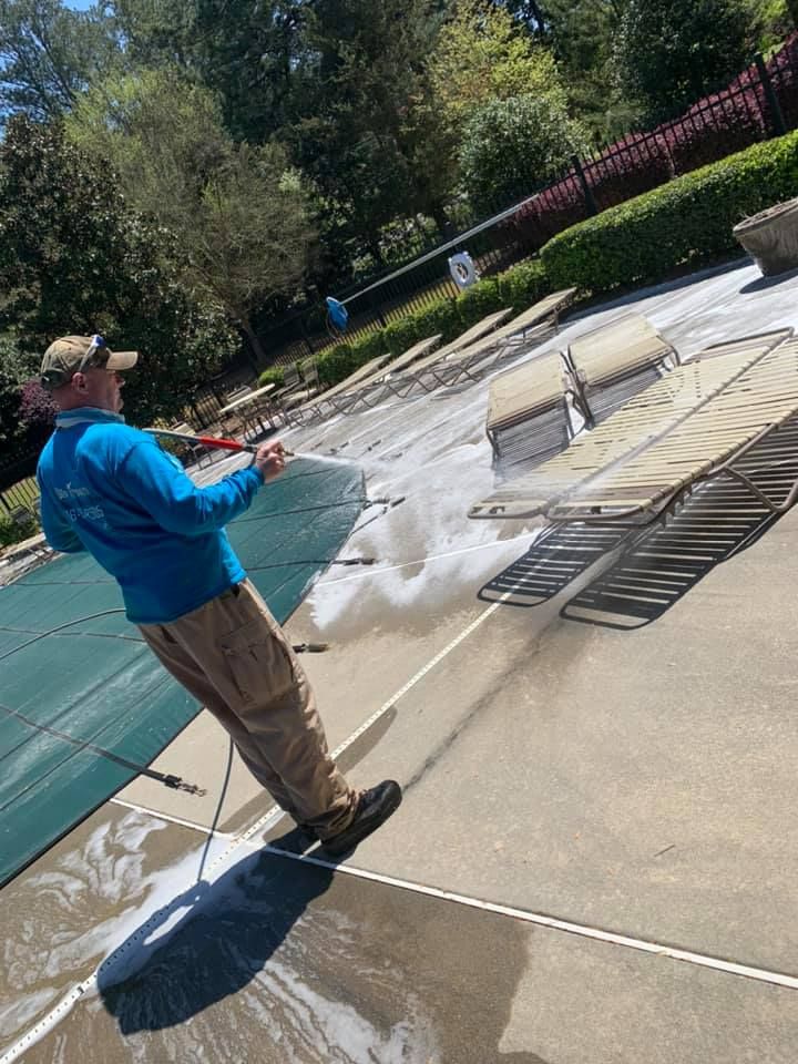 Person pressure-washes a pool deck beside covered lounge chairs and a swimming pool.