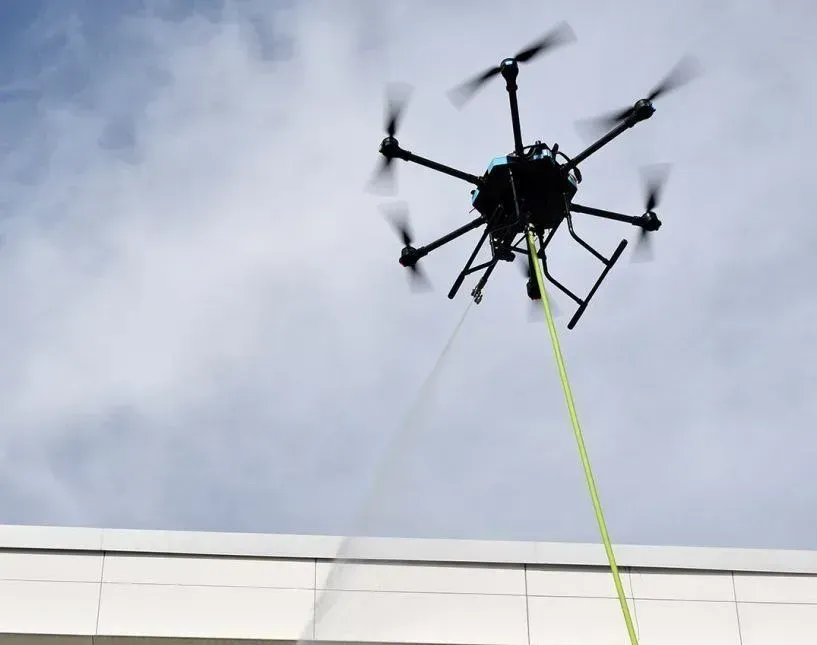 Drone hovering above a building, lowering a neon yellow rope against a cloudy sky