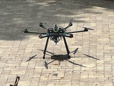 Large drone hovering over a sunlit stone-paved courtyard, casting a shadow below.
