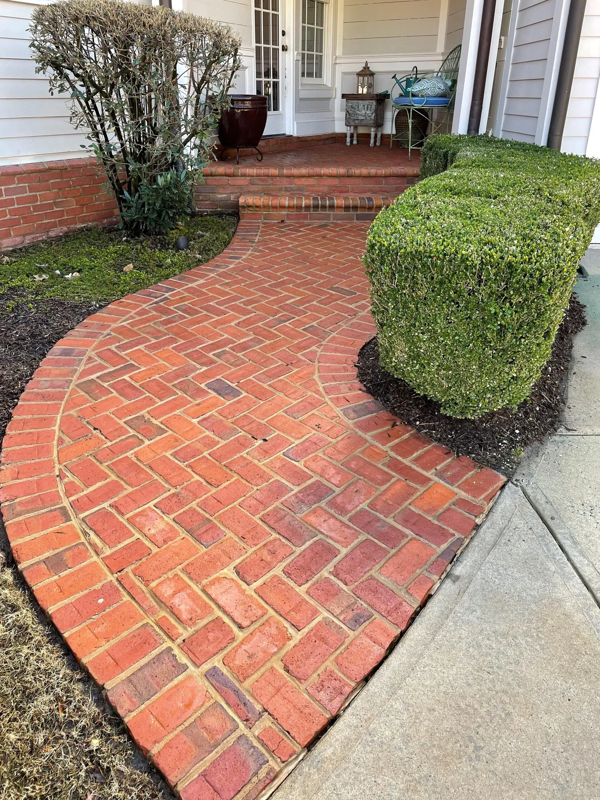 Curved red brick walkway leading to a front porch with trimmed hedges and concrete driveway edge