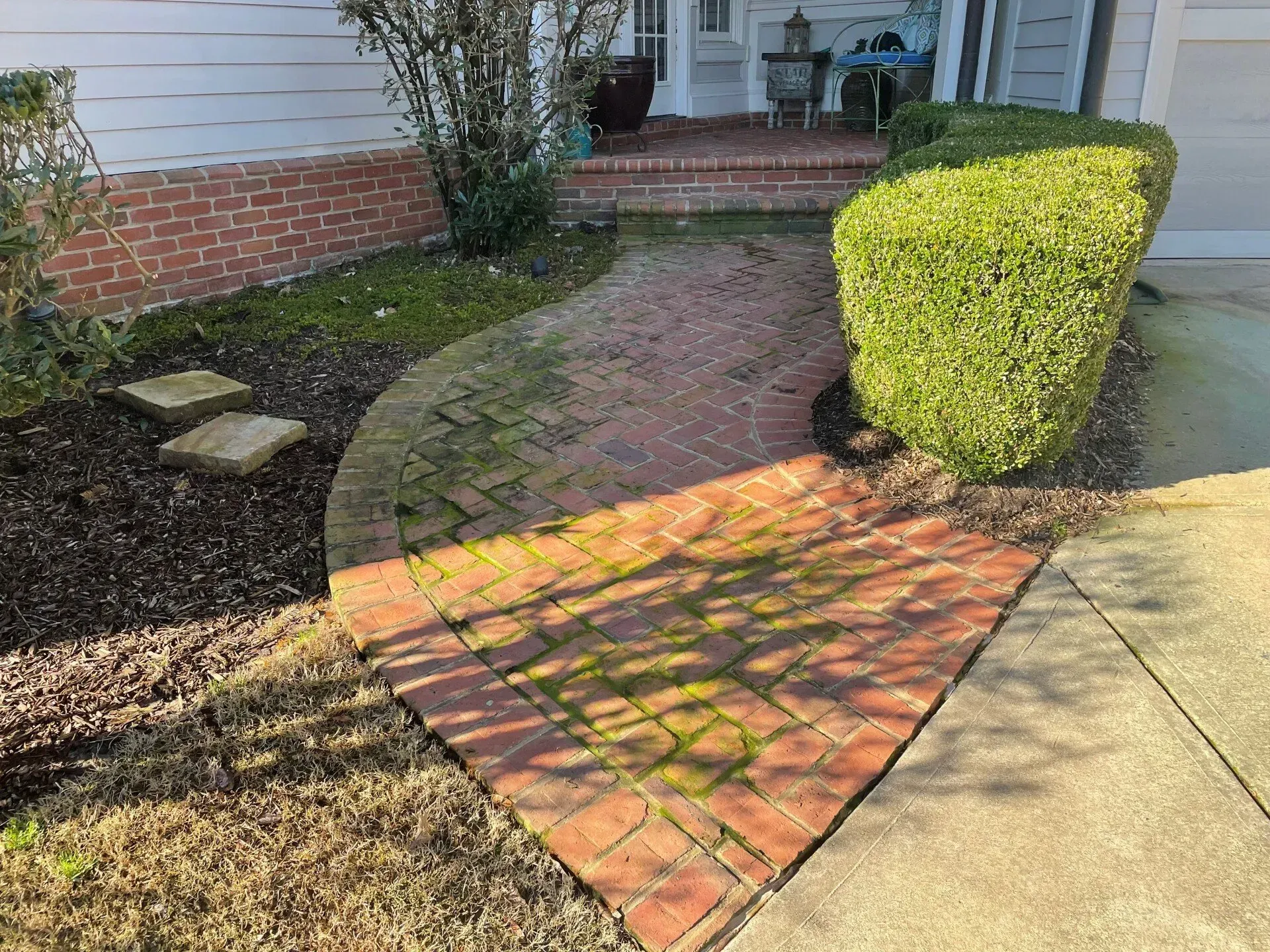 Brick walkway beside a house, curving past a trimmed shrub and lawn in sunlight.