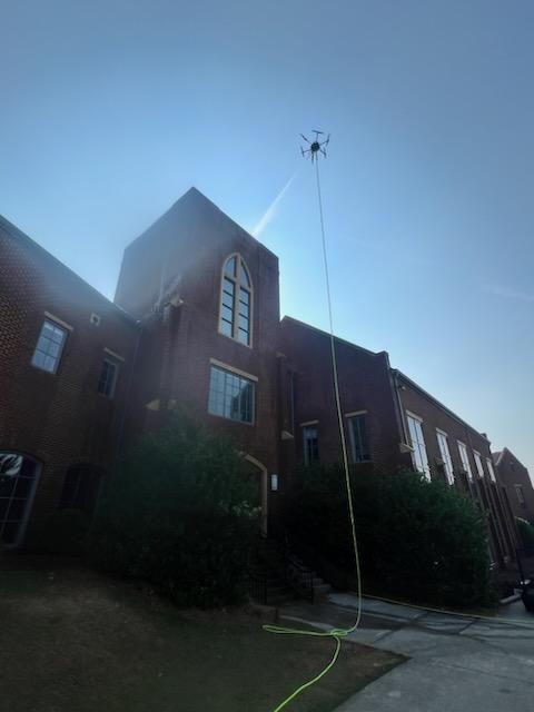 Brick building with a tall pointed roof and a hose spraying water upward in bright sunlight