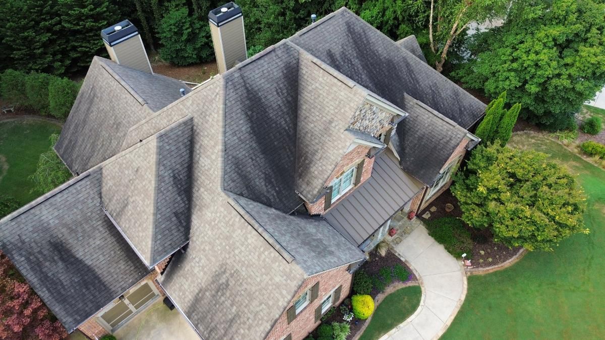 Aerial view of a brick house with a gray roof, chimneys, and surrounding green landscaping