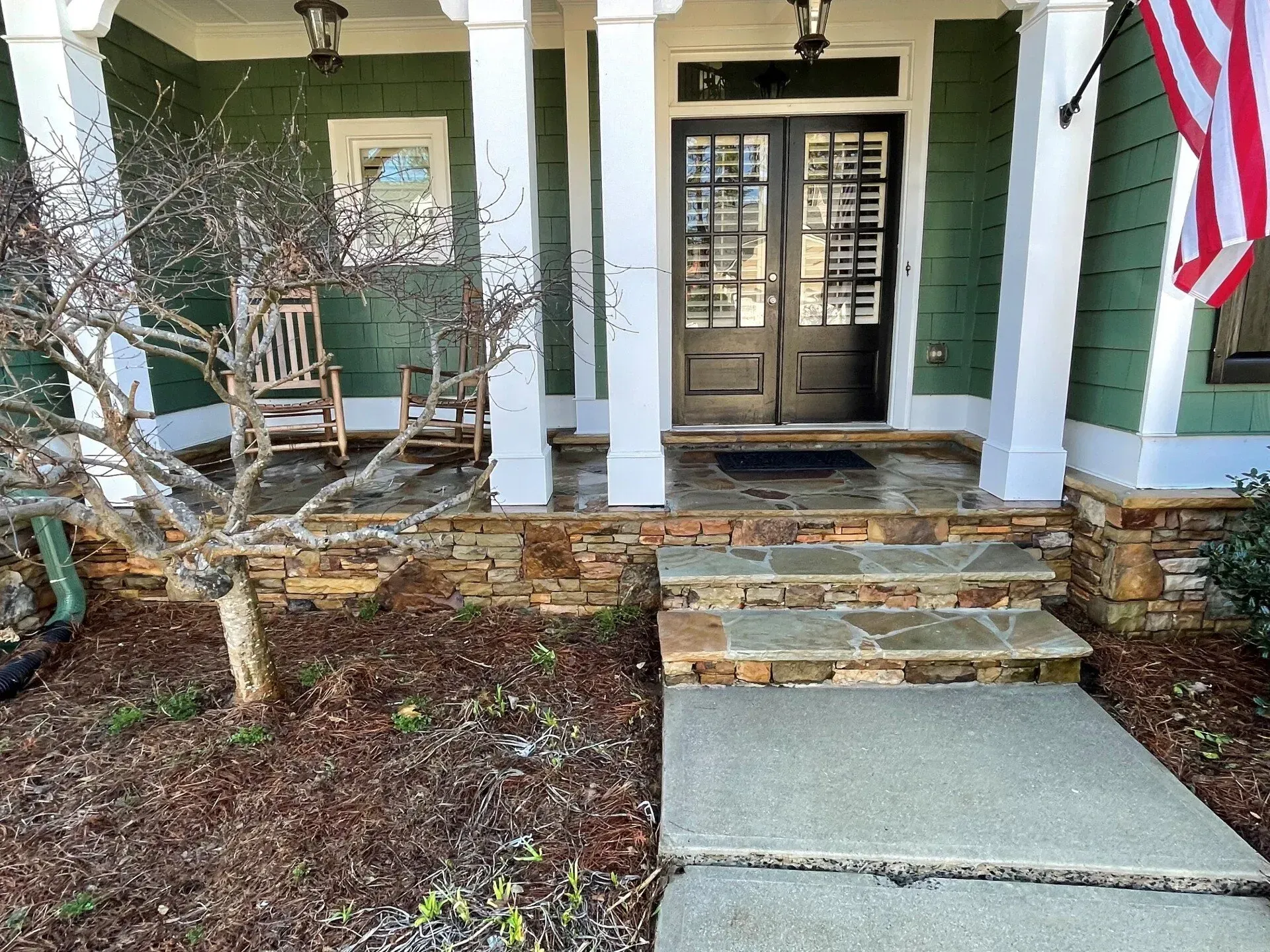 Front porch with stone steps, concrete ramp, white columns, green siding, and an American flag.