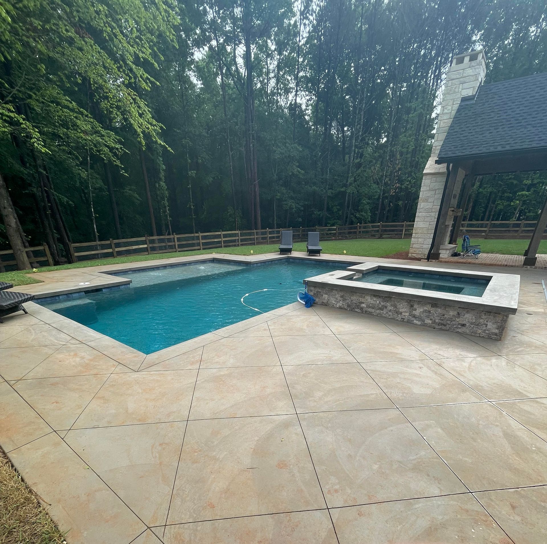 Backyard patio with a blue pool, stone hot tub, and surrounding trees and greenery