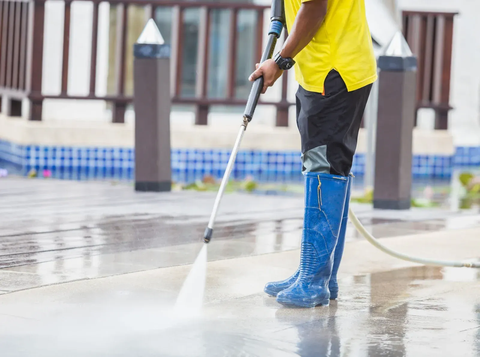 Person in blue boots, yellow shirt power washing a wet concrete surface near a pool.