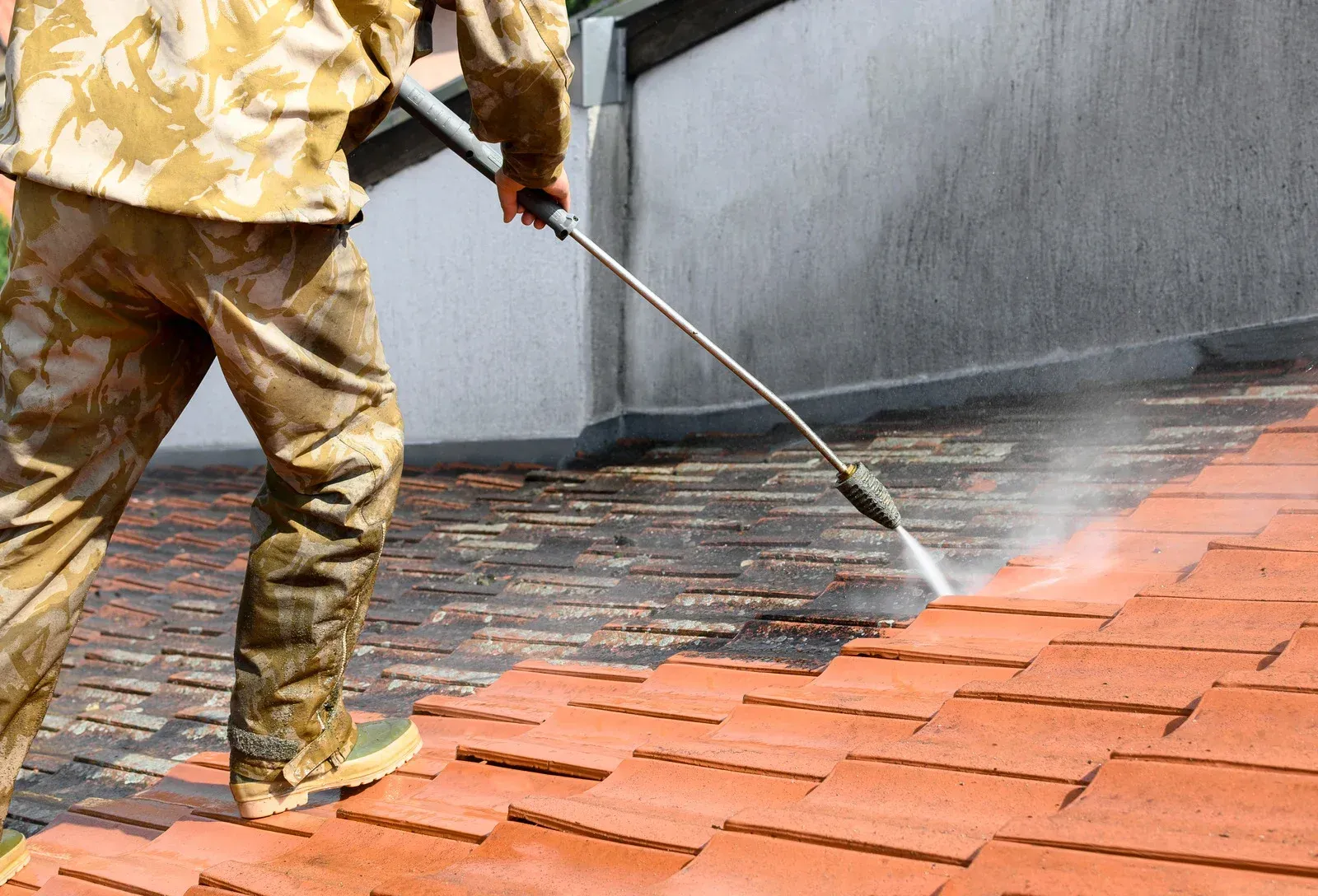 Worker pressure-washing orange roof tiles beside a gray wall